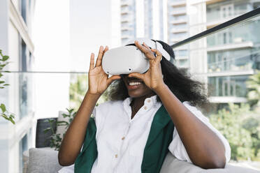 Glückliche Frau mit Virtual-Reality-Simulatoren auf dem Balkon sitzend - TYF00572
