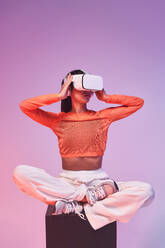 Junge Frau in modischem Outfit berührt VR-Headset und schaut weg, während sie im Schneidersitz auf einem Würfel sitzt und den Cyberspace vor violettem Hintergrund erkundet - ADSF42392