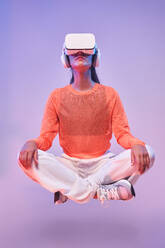 Ruhige Frau in stilvoller Kleidung mit VR-Brille schwebend in Lotus-Pose und meditierend vor violettem Hintergrund - ADSF42393