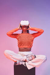 Junge Frau in modischem Outfit berührt VR-Headset und schaut weg, während sie im Schneidersitz auf einem Würfel sitzt und den Cyberspace vor violettem Hintergrund erkundet - ADSF42395