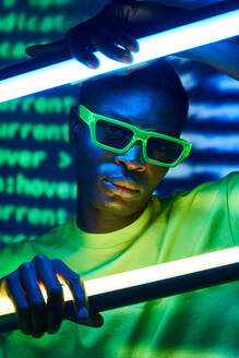 Emotionsloser afroamerikanischer Mann mit modischer Brille, der in die Kamera schaut, während er sich auf leuchtende, helle Leuchtstofflampen in der Nähe leuchtender Buchstaben stützt - ADSF42426