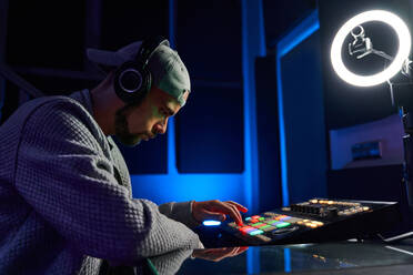 Side view of bearded man in cap sitting at workplace pushing button on mixer console while recording music in studio - ADSF42430