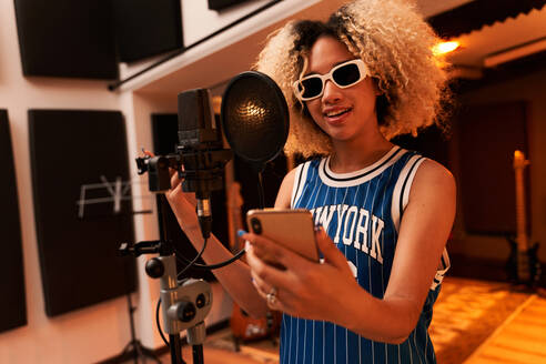 Positiver junger Musiker mit Afrofrisur und Sonnenbrille, der in einem Studio in der Nähe des Mikrofons steht und ein Lied singt, während er Musik aufnimmt und einen Text auf einem Mobiltelefon liest - ADSF42433