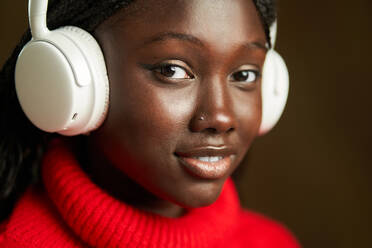 Seitenansicht einer positiven jungen schwarzen Frau in einem roten gehäkelten langärmeligen Oberteil, die mit Kopfhörern auf einem unscharfen braunen Hintergrund Musik hört - ADSF42448