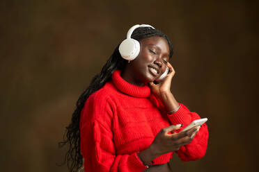 Seitenansicht einer positiven jungen schwarzen Frau in einem roten gehäkelten langärmeligen Oberteil, die ein Smartphone benutzt, während sie über Kopfhörer auf einem unscharfen braunen Hintergrund mit geschlossenen Augen Musik hört - ADSF42449
