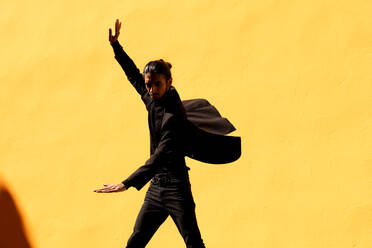 Hispanischer Mann im schwarzen Anzug mit Haarknoten gestikuliert und tanzt Flamenco gegen eine gelbe Wand - ADSF42456