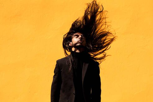 Young Hispanic male in black suit closing eyes and shaking long dark hair against yellow background - ADSF42462