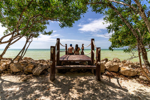 Rückenansicht eines Mannes und einer Freundin, die zusammen auf einem hölzernen Steg gegen das wogende Meer an einem sonnigen Tag in einem mexikanischen Ferienort sitzen - ADSF42474