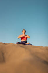 Full body of young woman in sportswear sitting on sandy beach in lotus pose and meditating with closed eyes - ADSF42479