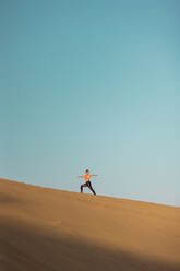 Fernblick auf eine junge Frau, die auf einer Sanddüne in der Wüste Yoga übt. Kriegerhaltung. - ADSF42481