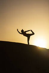 Seitenansicht der Silhouette einer anonymen Frau, die am Rande eines Berges steht und die Arme hebt, während sie am Abend Yoga übt - ADSF42482
