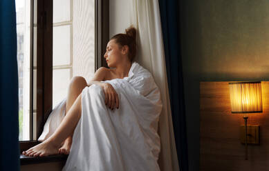 Carefree woman relaxing next to the window and being wrapped in blanket - ADSF42497