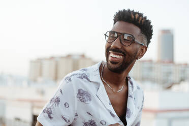 Positiv lächelnder schwarzer Mann in Hemd, Jeans und Brille mit Armbanduhr auf einem Betonbalkon, während er auf einem hohen Stockwerk eines Gebäudes mit Blick auf die Stadt steht - ADSF42507