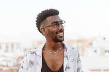 Positive smiling black male in shirt jeans and eyeglasses with wristwatch on concrete balcony while standing looking away on high floor of building with view on city - ADSF42509