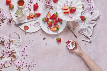 Draufsicht auf eine nicht erkennbare Frau mit einem Becher köstlichen Joghurts an einem Tisch mit Tellern mit reifen frischen Erdbeeren und Krügen, die mit Zweigen des Sakura-Baums geschmückt sind - ADSF42515