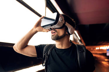 Young bearded male with backpack in black t shirt and VR goggles experiencing virtual reality while standing on street near wall with illuminated lights - ADSF42532