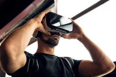 Young bearded male with backpack in black t shirt and VR goggles experiencing virtual reality while standing on street near wall with illuminated lights - ADSF42533