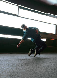 Ganzer Körper eines Mannes in Freizeitkleidung, der auf Zehenspitzen stehend auf einer asphaltierten Straße tanzt, in der Nähe einer modernen Wand mit leuchtenden Lampen - ADSF42534