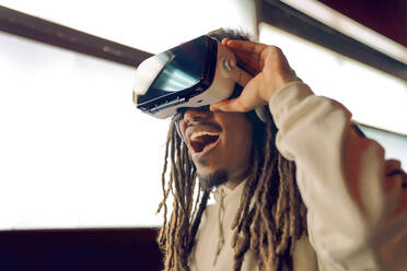 Junger staunender Afroamerikaner, der eine VR-Brille berührt und virtuelle Realität in einem modernen Raum mit leuchtenden Lampen erlebt - ADSF42535