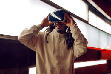 Junger glücklicher afroamerikanischer Mann, der eine VR-Brille berührt und virtuelle Realität in einem modernen Raum mit leuchtenden Lampen erlebt - ADSF42537