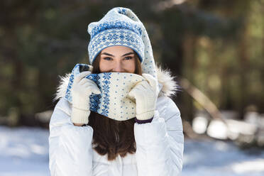 Happy lady in winter clothes looking at camera and touching long tip of cap while standing in snowy forest in sunlight - ADSF42538