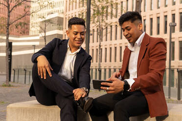 Lächelnde junge ethnische Männer in stilvoller Kleidung, die am Zaun sitzen und mit dem Handy im Internet surfen, während sie sich im Freien ausruhen - ADSF42539