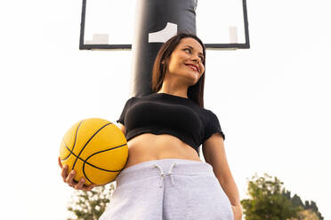 Low angle of cheerful young female in sportswear standing near hoop with yellow ball during break from streetball game looking away - ADSF42546