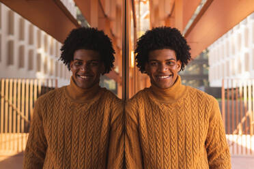 Optimistischer afroamerikanischer Mann mit Afrohaar in gelbem Pullover, der lächelt und in die Kamera schaut, in der Nähe eines Spiegels mit Reflexion auf der Terrasse eines modernen Gebäudes - ADSF42550