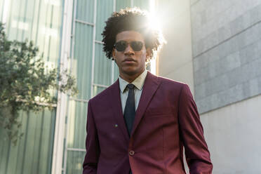 Selbstbewusster afroamerikanischer Mann im eleganten bordeauxfarbenen Anzug und mit Sonnenbrille und lockigem Haar, der die Hand in der Tasche hält und in die Kamera schaut, während er in der Nähe eines modernen Glasgebäudes auf einer Stadtstraße steht - ADSF42558