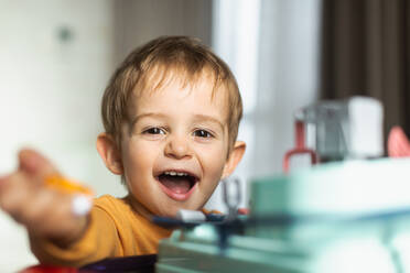 Fröhlicher, liebenswerter, kleiner, blondhaariger Junge im orangefarbenen Pullover, der am Tisch sitzt und mit Spielzeug spielt, während er Zeit zu Hause verbringt - ADSF42563