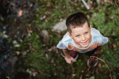 Weichzeichner Draufsicht eines fröhlichen Jungen in Freizeitkleidung, der lächelt und in die Kamera schaut, während er einen Wochenendtag in der Natur verbringt - ADSF42579