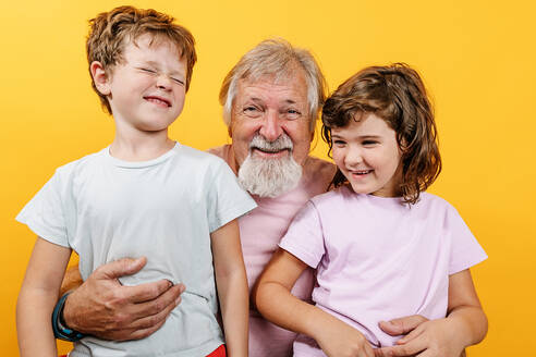 Fröhlicher grauhaariger älterer Mann umarmt lachenden kleinen Jungen und Mädchen, während er im Studio steht - ADSF42587