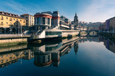 Malerische Kulisse von La Ribera Bilbao in der Nähe des ruhigen Flusses, in dem sich die Gebäude der Stadt unter blauem Himmel spiegeln, im Baskenland - ADSF42597