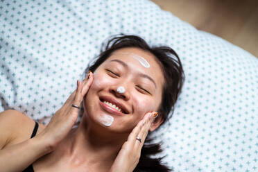 From above of ethnic female with eyes closed smiling and enjoying process of facial massage while spreading cream and lying - ADSF42601