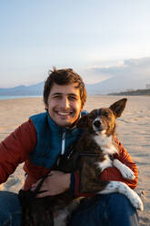 Glücklicher Mann in warmer Jacke umarmt niedlichen Stirn Hund mit weißen Flecken am Strand in der Nähe von Meer am sonnigen Tag mit blauen wolkenlosen Himmel Blick auf die Kamera - ADSF42632
