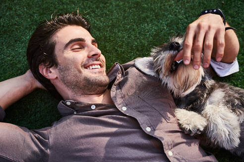Draufsicht auf einen erwachsenen bärtigen Mann im Hemd, der auf einer grünen Wiese liegt und einen süßen Hund streichelt, an einem sonnigen Tag - ADSF42641