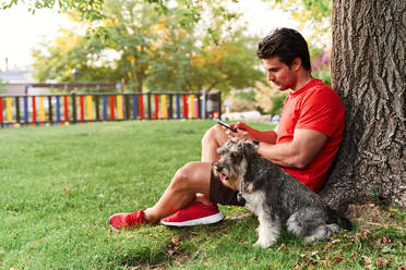 Side view full body of adult male athlete in red sportswear sitting near tree and texting message via smartphone near cute Miniature Schnauzer during walk in park - ADSF42648