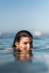 Frau mit dunklen nassen Haaren schwimmt im ruhigen Wasser des Meeres, während sie in die Kamera auf unscharfen Hintergrund des blauen Himmels schaut - ADSF42668
