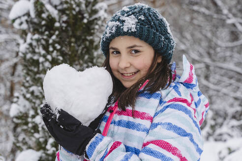 Smiling girl wearing warm clothing showing heart-shaped snowball - OSF01274