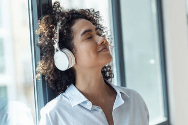 Lächelnde Frau mit drahtlosen Kopfhörern, die sich zu Hause ans Fenster lehnt - JSRF02332