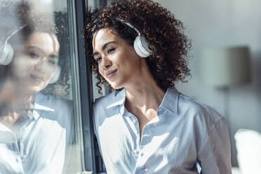 Contemplative smiling woman with headphones looking through window - JSRF02334