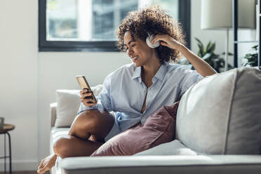 Smiling woman with headphones using smart phone on sofa at home - JSRF02339