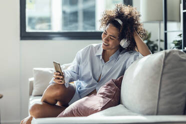 Happy woman using mobile phone sitting on sofa in living room - JSRF02340