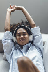 Young woman wearing headphones resting on sofa at home - JSRF02347