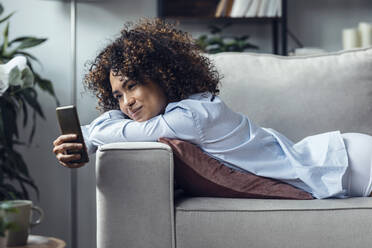 Young woman using mobile phone lying on sofa at home - JSRF02349