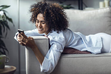 Happy woman text messaging over smart phone lying on sofa - JSRF02350