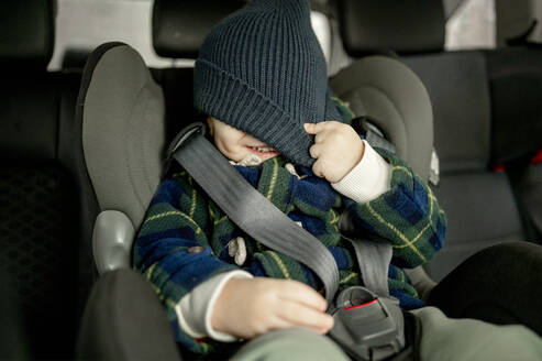 Glücklicher Junge, der sein Gesicht mit einer Strickmütze im Autositz bedeckt - ANAF00789