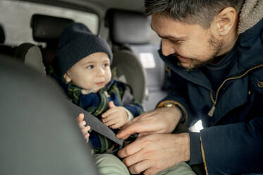 Lächelnder Vater, der den Sicherheitsgurt für seinen Sohn im Auto befestigt - ANAF00790