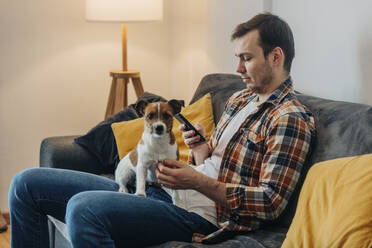 Mann mit Smartphone auf dem Sofa sitzend mit Hund zu Hause - VSNF00261