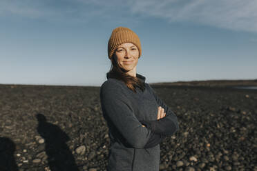 Happy woman wearing knit hat standing with arms crossed at Janubio Beach, Lanzarote, Canary Islands, Spain - DMGF00930